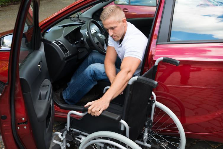 Person sitting in driver's seat of a red car with door open and wheelchair beside them.