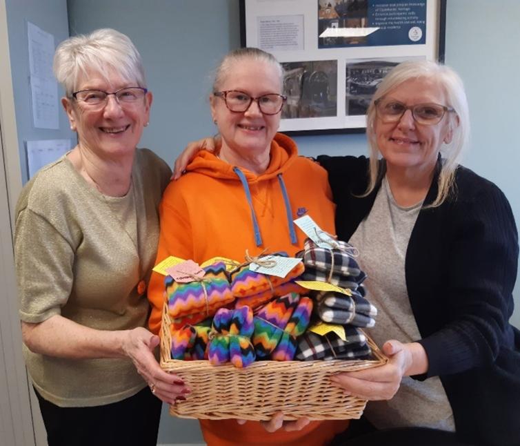 Three people all stood together and smiling. Between them they are holding a woven brown basket. Inside the basket are multi-coloured handwarmers.
