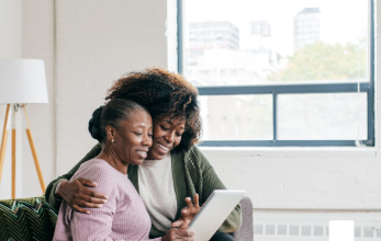 Two people sat next to each other on a sofa. Both are smiling at a tablet.