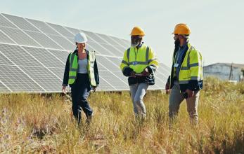 Three people stood in a field with ground mounted solar panels, all wearing hard hats and high visibility jackets. 