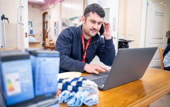 A person wearing a blue jumper and red lanyard, sat at a table in a community space. They are talking into a mobile phone. On the table is a laptop and boxes of LED light bulbs.