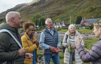 A small group of adults standing on the shore of a sea loch. They are all wearing warm clothing and looking at one of the group members who is talking. The shoreline is rocky and there are a couple of houses and rugged hills behind them.