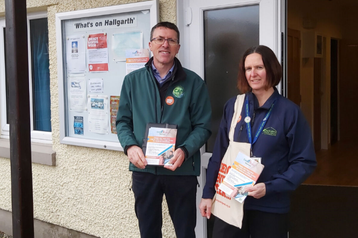 Two people stand outside a building entrance holding leaflets and a tote bag. A noticeboard with notices is mounted on the building wall behind them.