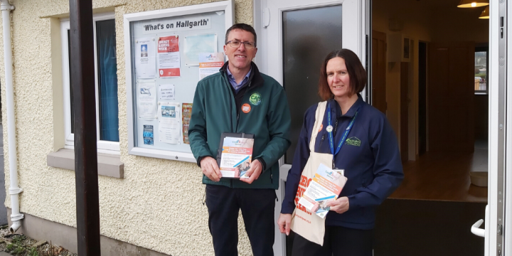Two people stand outside a building entrance holding leaflets and a tote bag. A noticeboard with notices is mounted on the building wall behind them.