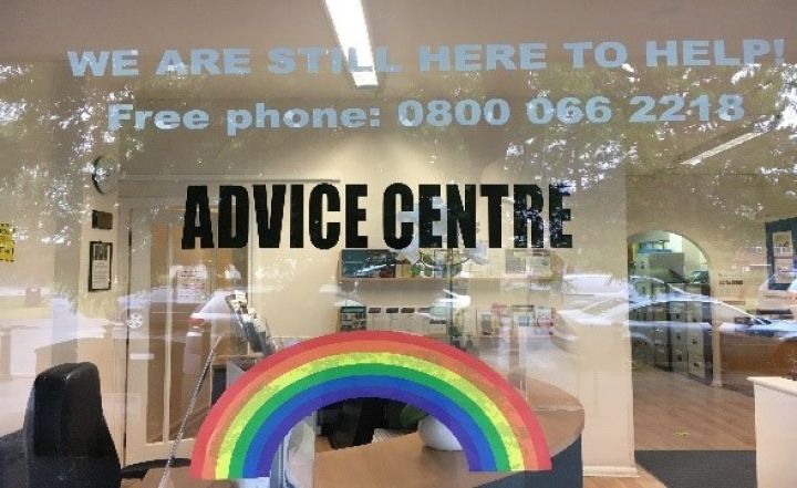 Advice Centre window with a rainbow, contact number, and WE ARE STILL HERE TO HELP! message; office interior and parked cars visible through the glass.