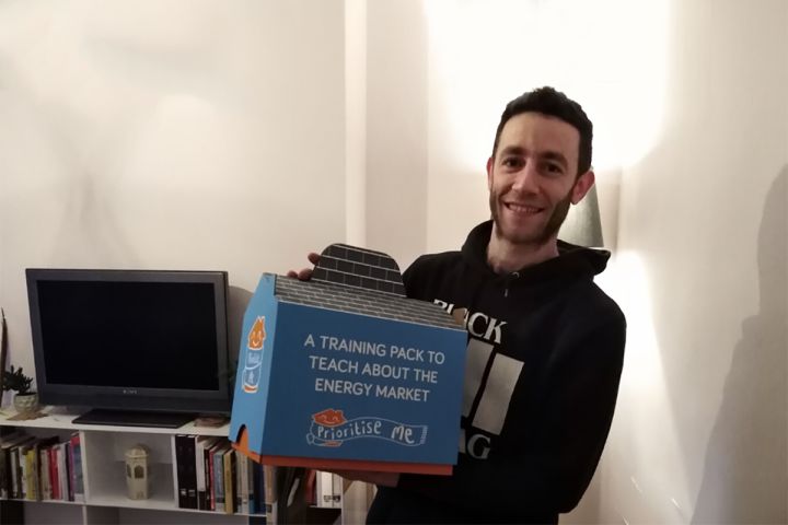 A person holding a blue box labelled A TRAINING PACK TO TEACH ABOUT THE ENERGY MARKET whilst standing in a living room with a TV and bookcase.