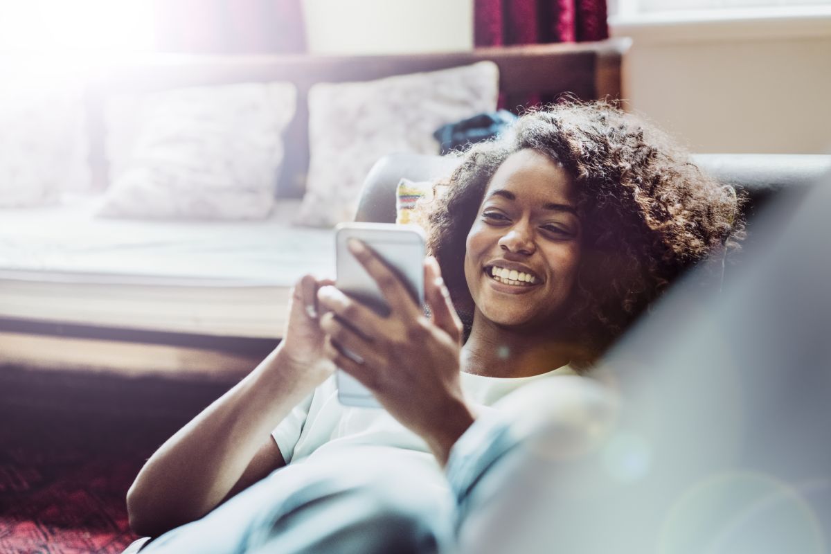 A person is lying on a sofa, smiling whilst looking at a mobile phone. There are cushions and sunlight in the background.