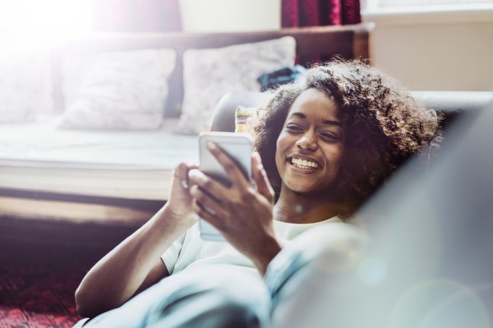 A person is lying on a sofa, smiling whilst looking at a mobile phone. There are cushions and sunlight in the background.