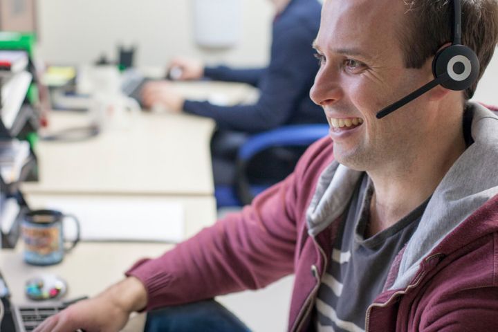 a person wearing a headset sits at a desk, smiling and looking at a computer screen. Another person works at a computer in the background.