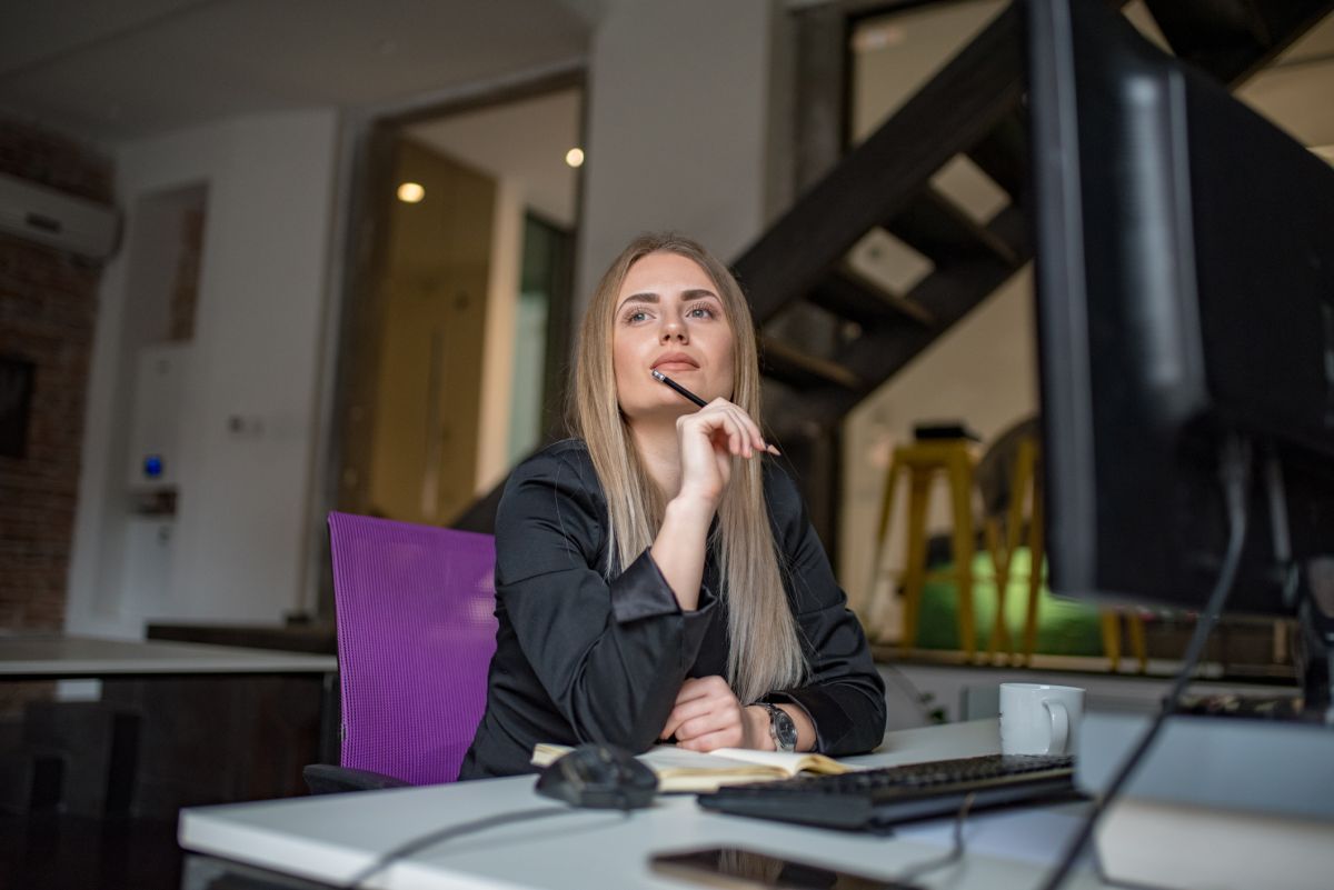 A woman sits at a desk in front of a computer, holding a pen to her chin and looking thoughtful.