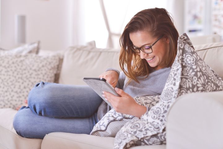 A woman wearing glasses sits on a sofa, smiling and using a tablet. She is leaning on patterned cushions in a bright, cosy living room.