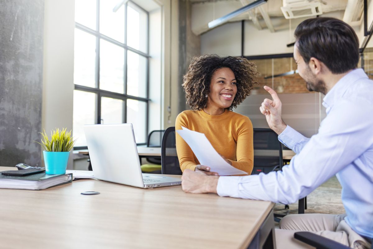 Two colleagues sit at a desk in a modern office, talking. One holds papers and smiles whilst the other gestures with a raised finger. A laptop and plant are on the desk.