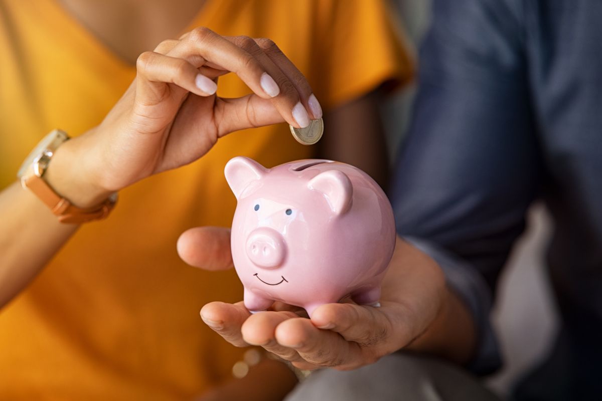 A person holds a pink piggy bank while another hand drops a coin into it. Both people are partially visible, wearing casual clothes.