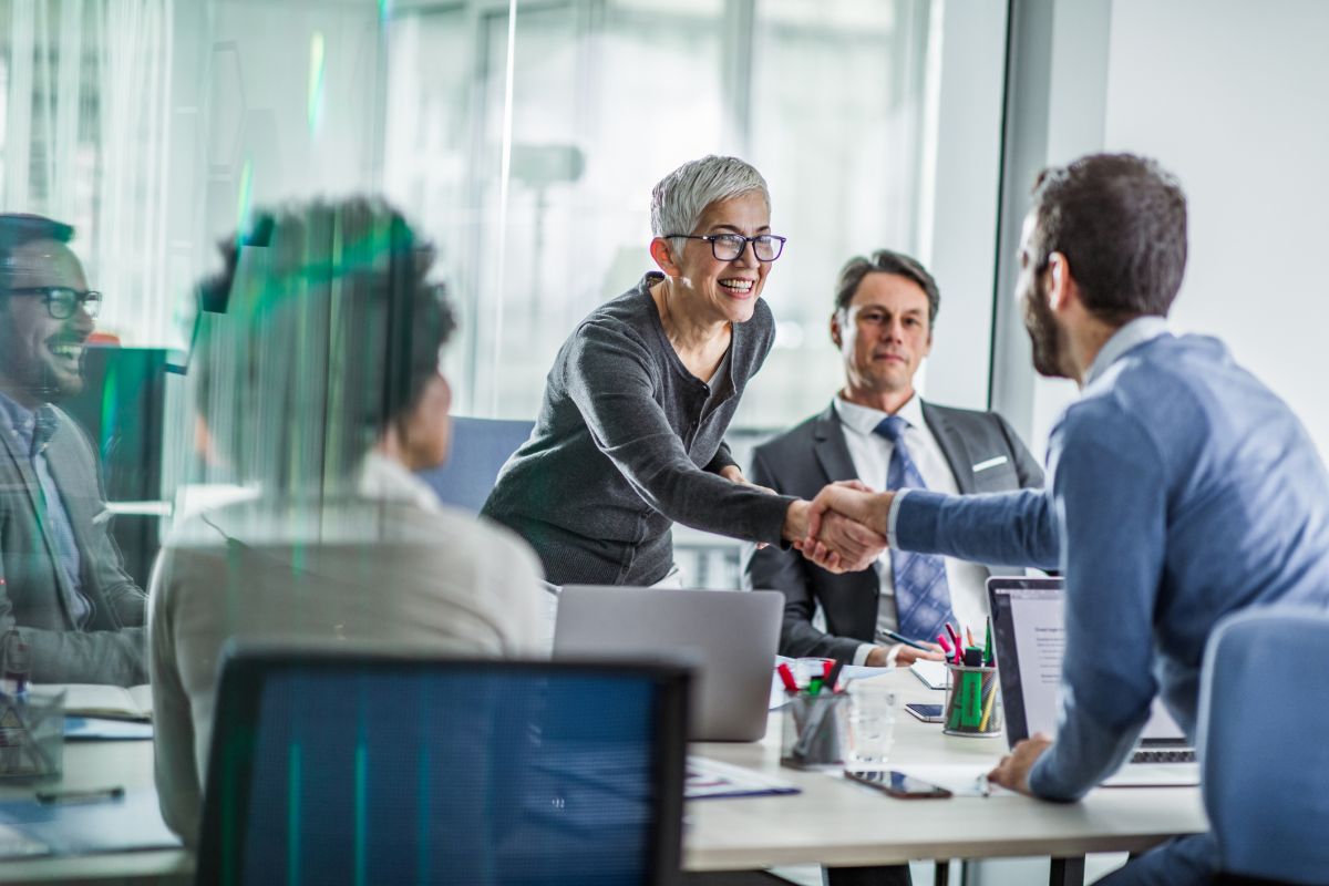 A woman stands and shakes hands with a seated man in a modern office meeting room whilst three other people watch.