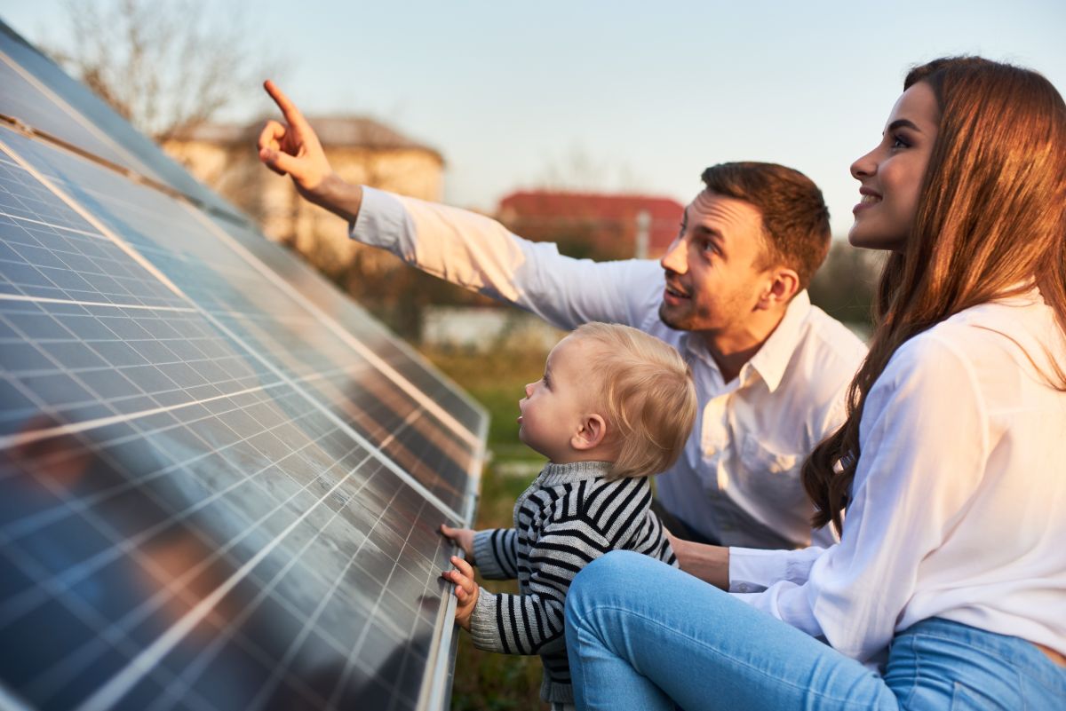 A man, woman, and young child look at and touch a solar panel outdoors, with the man pointing towards something in the distance.