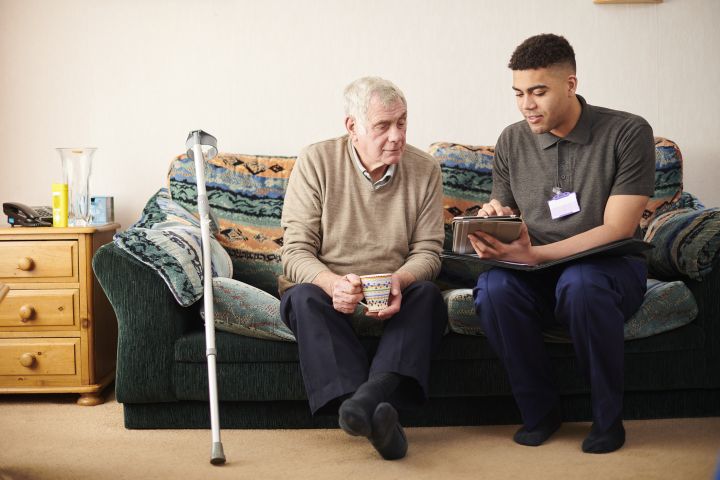 A young carer shows a tablet to an older man sitting on a sofa, who is holding a mug. A crutch leans against the sofa.