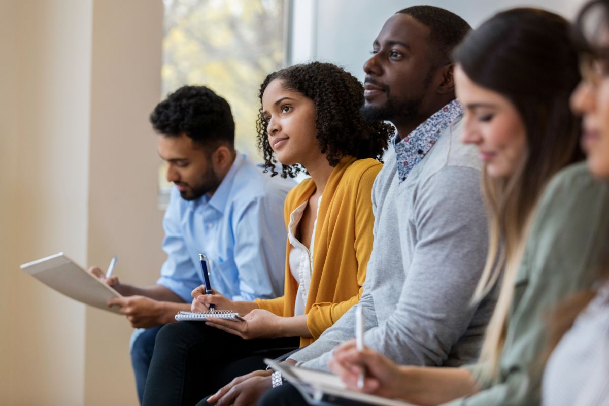 Four adults sit in a row, taking notes in notebooks, appearing focused and attentive during what looks like a meeting or class.