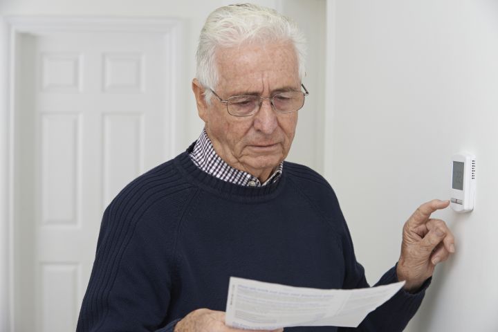 Older man adjusts a wall-mounted thermostat while reading a piece of paper, possibly instructions or a bill, in a home setting with a white wall and door in the background.