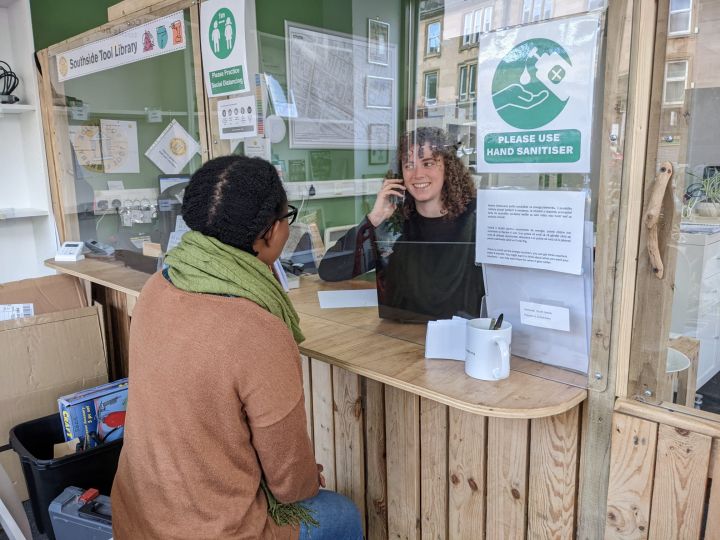 A person sits at a wooden counter speaking to a member of staff behind a glass screen in an office. Notices about hand sanitiser and hygiene are posted on the glass.
