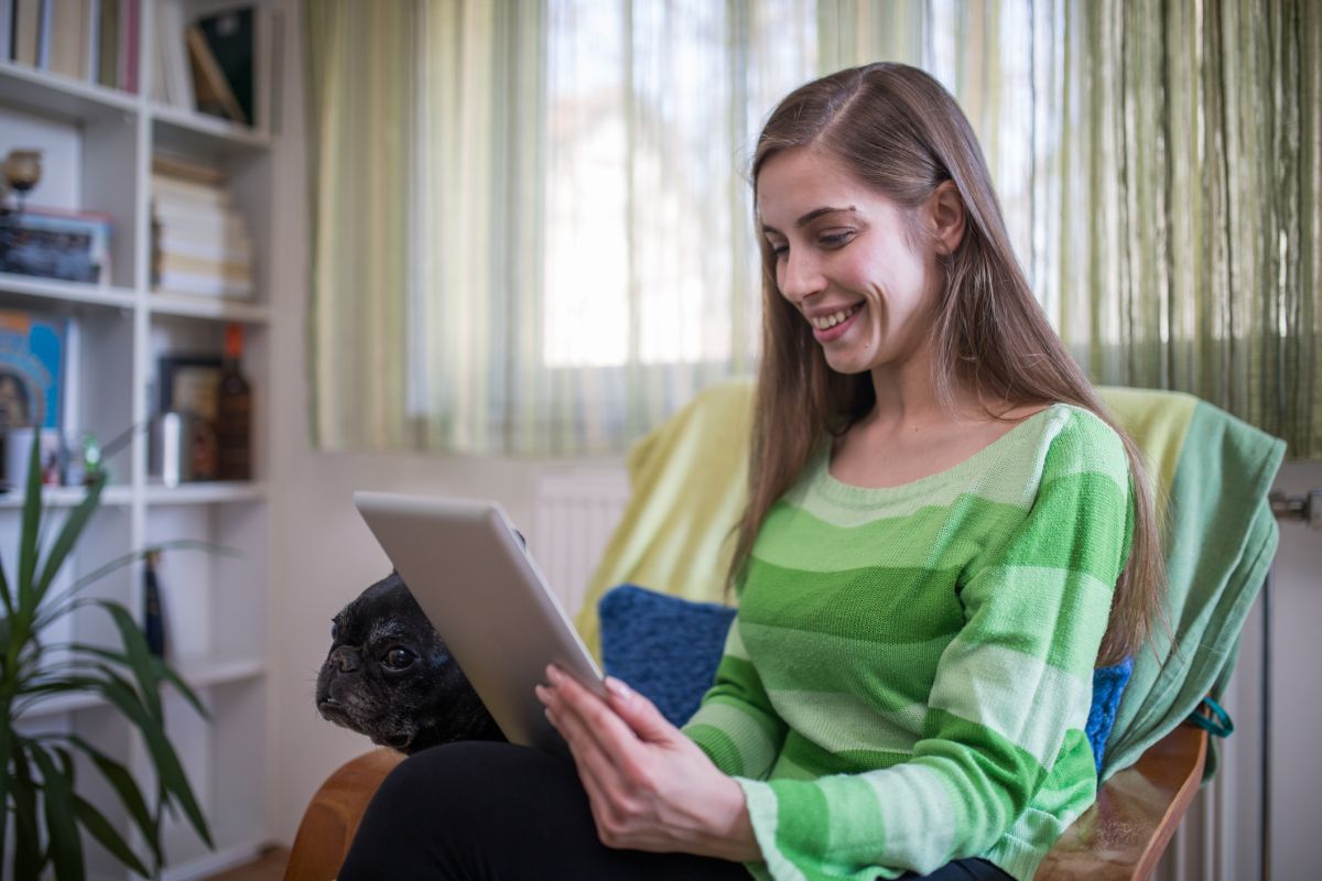 A woman in a green striped jumper sits indoors, smiling at a tablet. A black dog sits next to her. Shelves and a window with curtains are in the background.
