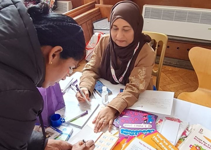 A woman in a brown hijab sits at a table with papers and colourful materials while another woman leans over to fill in a form.