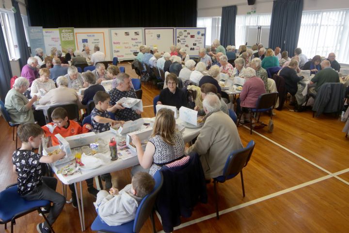 A group of people, mostly elderly, sit at tables in a hall eating meals. Several children are present. Posters are displayed on a board at the front. The room has wooden floors and blue chairs.