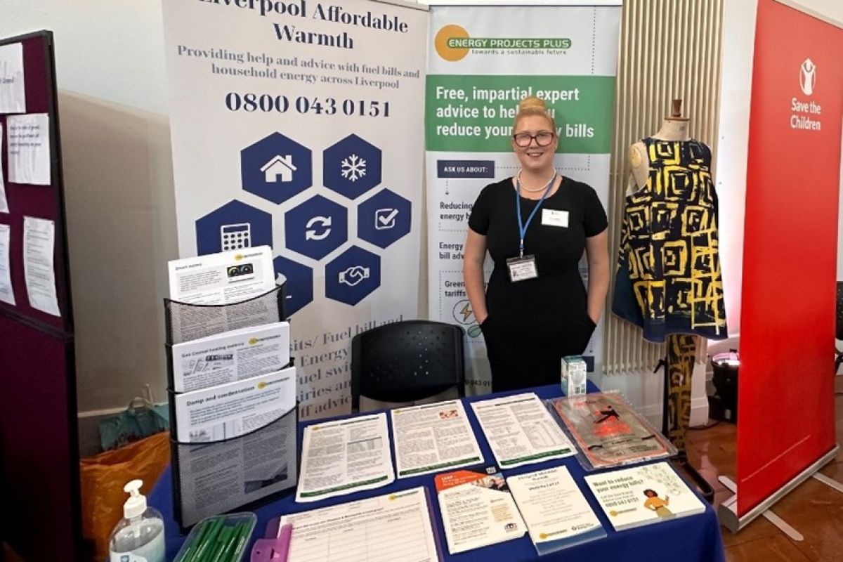 A woman stands behind a table with informational materials and leaflets at an indoor event stand for Liverpool Affordable Warmth and Energy Projects Plus.