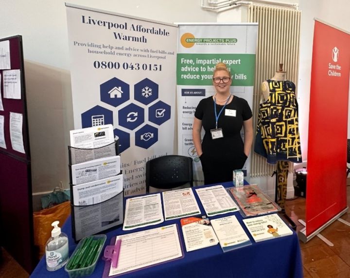 A woman stands behind a table with informational materials and leaflets at an indoor event stand for Liverpool Affordable Warmth and Energy Projects Plus.