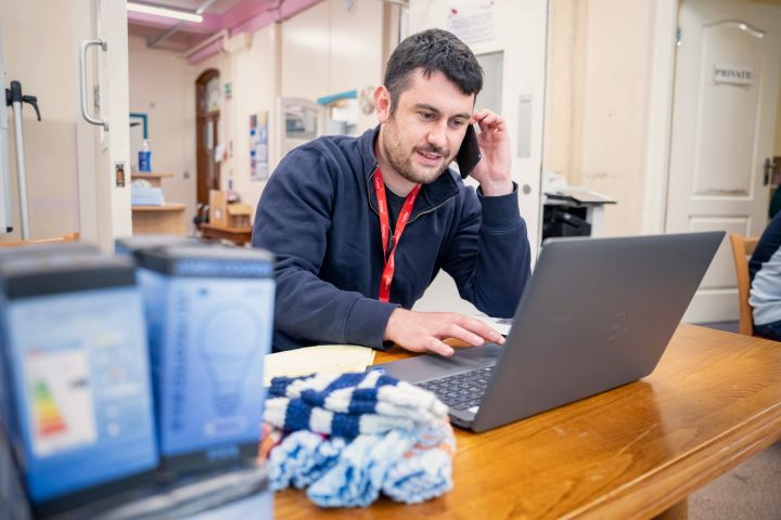 man with a red lanyard talks on the phone while typing on a laptop in a workspace