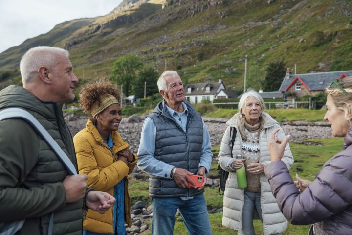 small group of adults wearing warm walking clothing gathered and talking as a group with a couple of houses and rugged hills behind them
