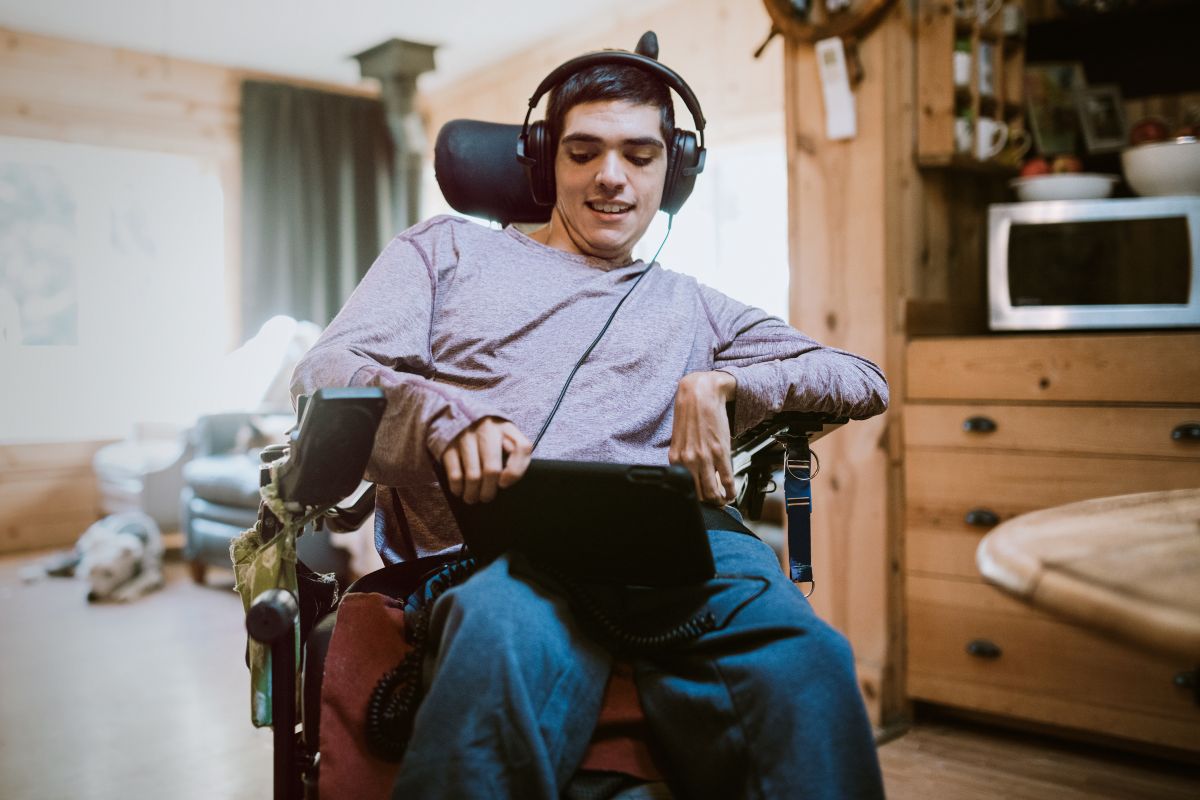 young adult man with cerebral palsy in a wheelchair at home listens to or watches something on a digital touchscreen tablet