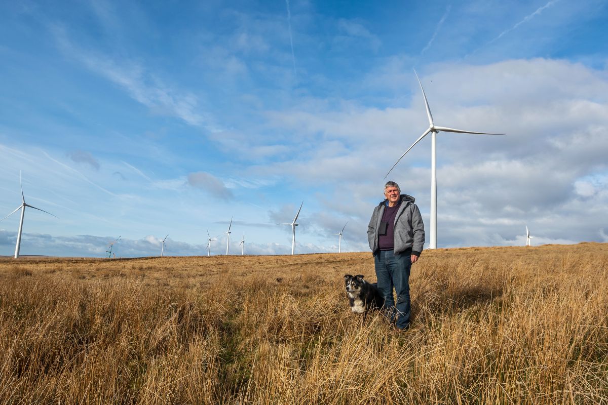senior man in a hooded jacket stands with a dog on Welsh moorland with wind turbines in the distance