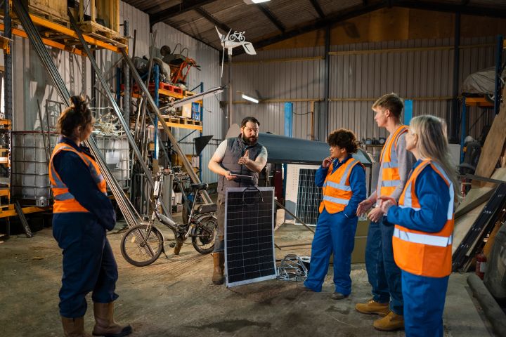 young team wearing hi-vis jackets and work overalls standing in a group listening to a man holding a solar panel in a workshop filled with electric bikes and solar panels