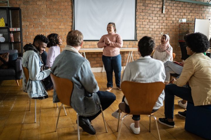 young woman talking to a relaxed group sat in chairs at a community meeting