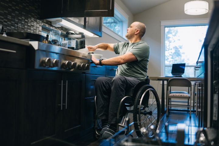 man using a wheelchair puts dishes away in the cabinets of his kitchen