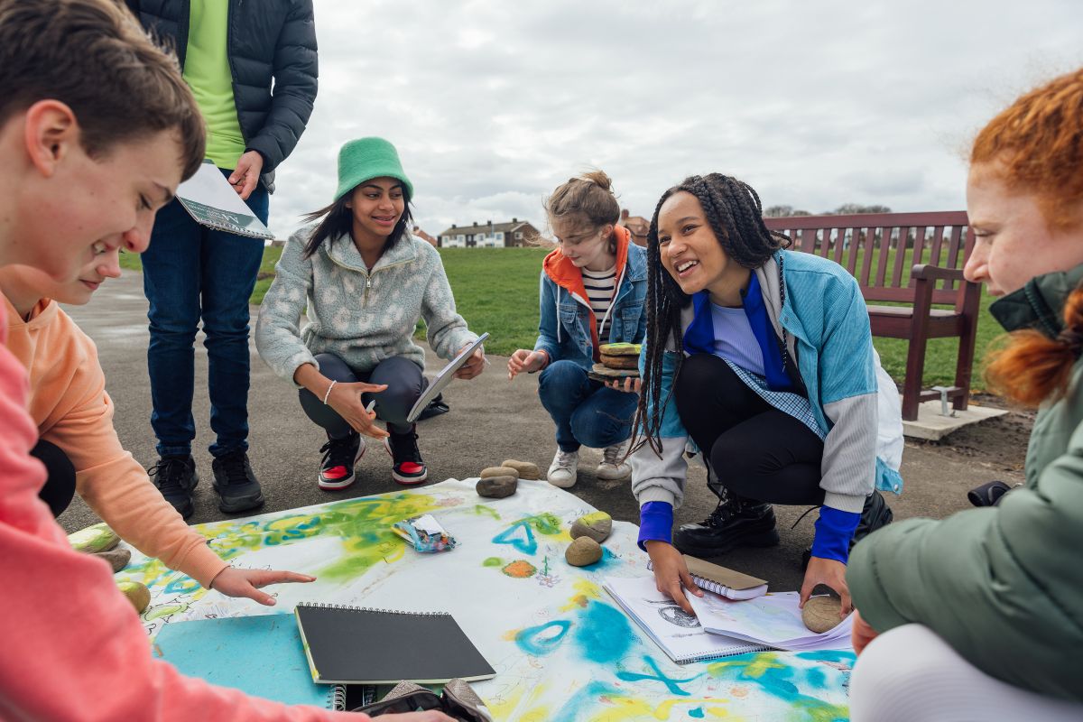 a group of teenagers wearing casual clothing on an overcast summer day, gathered near a park bench and using different arts and crafts products to create a sustainability artwork