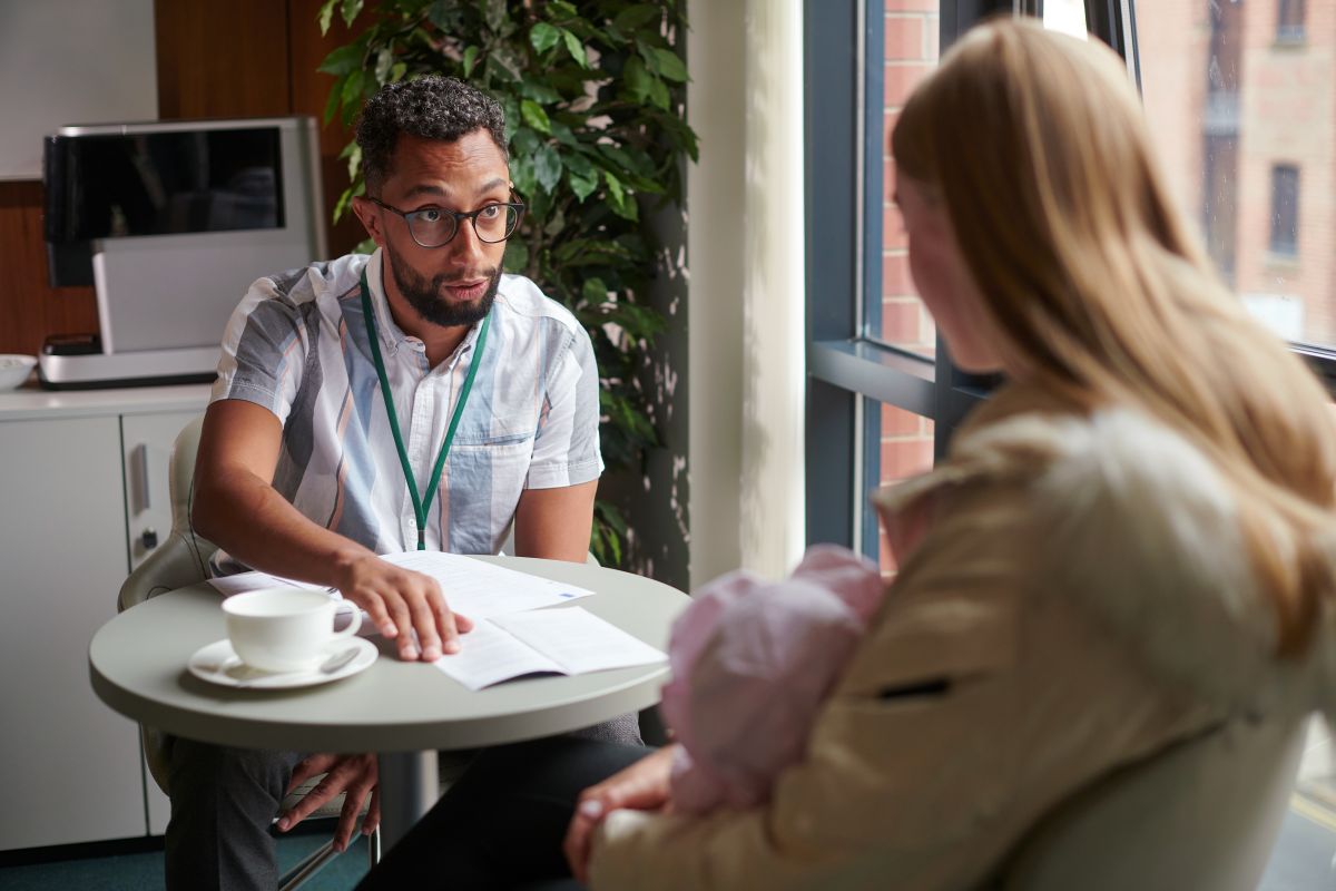 young mum getting help from a man wearing a lanyard, they are sitting at a small table with paperwork and a coffee cup