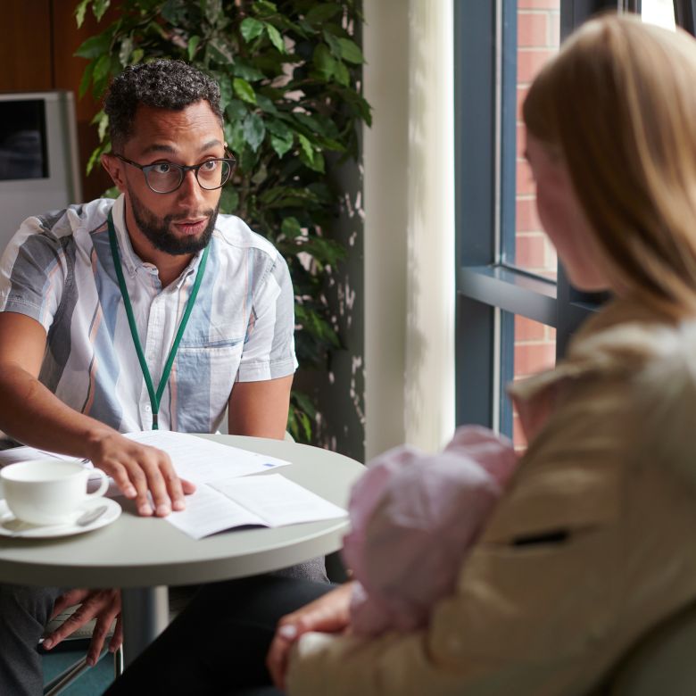 young mum getting help from a man wearing a lanyard, they are sitting at a small table with paperwork and a coffee cup