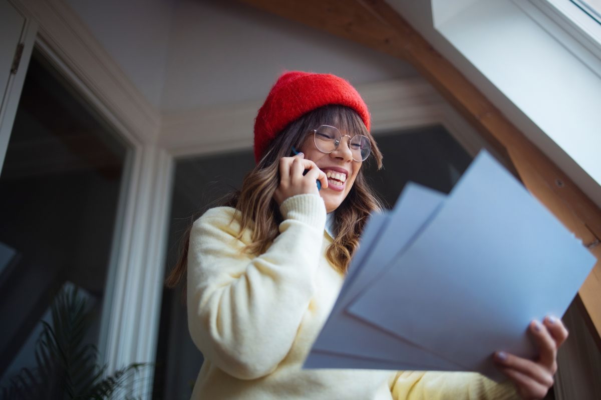 a cheerful young woman wearing a red hat talking on the phone and holding papers in a bright indoor setting