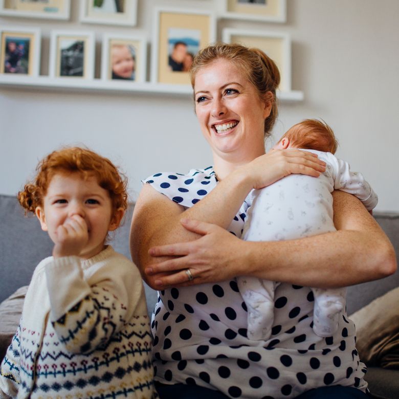 smiling mother patting a young baby over her shoulder with a toddler next to her on a sofa at home
